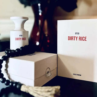 Imagen del perfume Borntostandout Dirty Rice. Botella blanca con etiqueta roja y caja minimalista, acompañada de una caja abierta y cuentas negras sobre fondo oscuro.