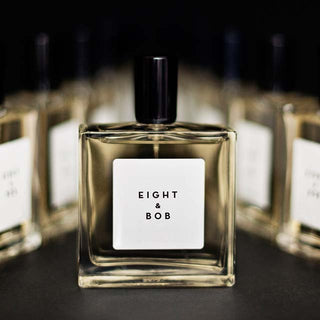 Primer plano de una botella de perfume Eight & Bob con etiqueta blanca y texto negro, elegantemente cuadrada con un tapón oscuro. Múltiples botellas idénticas se difuminan en el fondo.