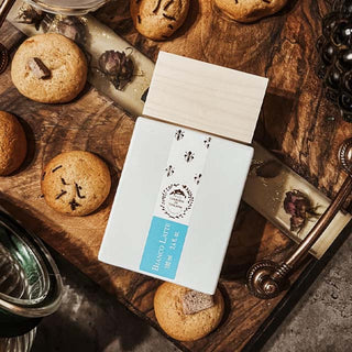 Imagen estilizada del perfume Bianco Latte de Giardini di Toscana: botella blanca de Eau de Parfum en caja con etiqueta azul, rodeada de galletas con chispas de chocolate sobre una tabla de madera rústica, rosas marchitas y un toque de vapor lechoso.